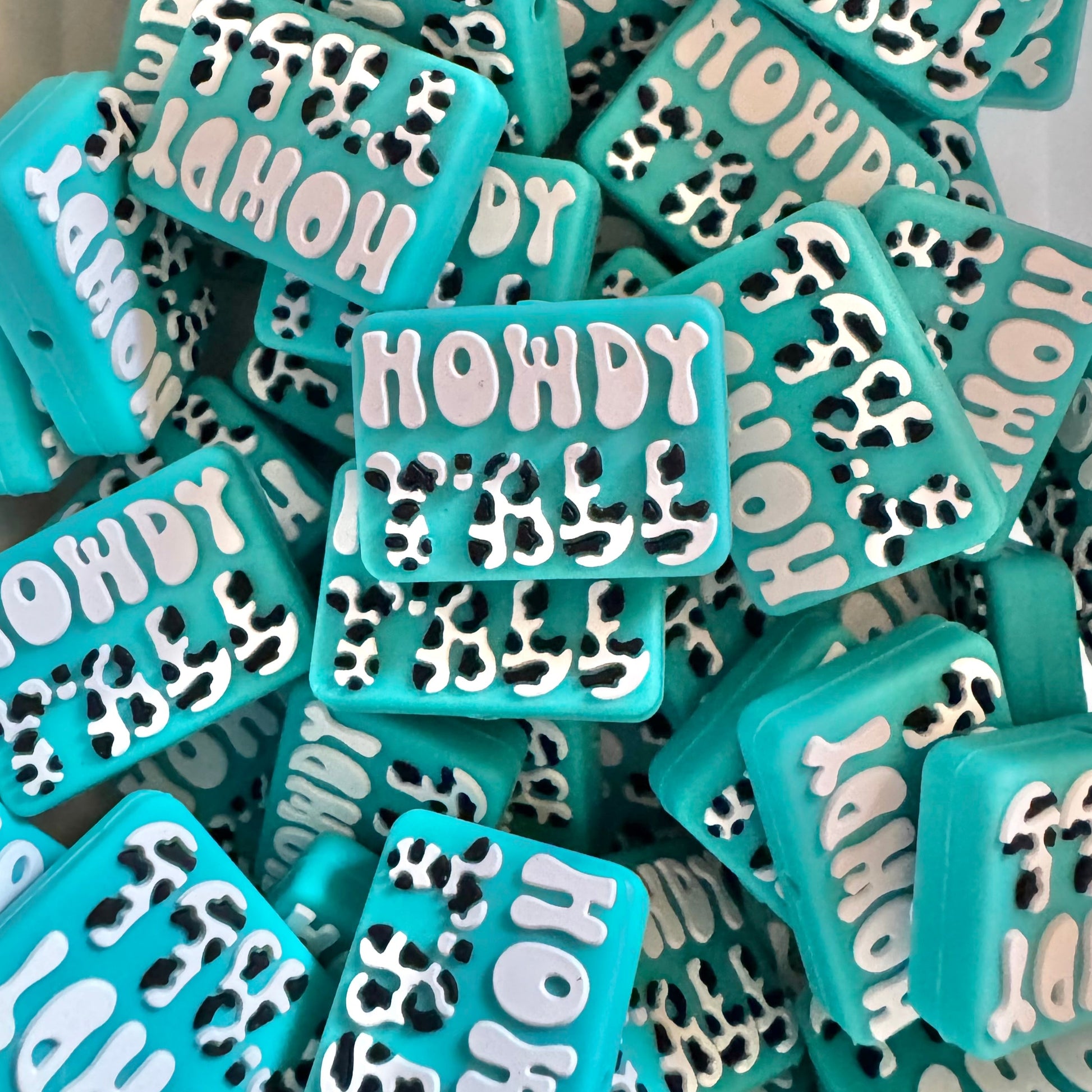 Turquoise rectangular beads with 'HOWDY FOLLE' text and leopard print design on a white surface